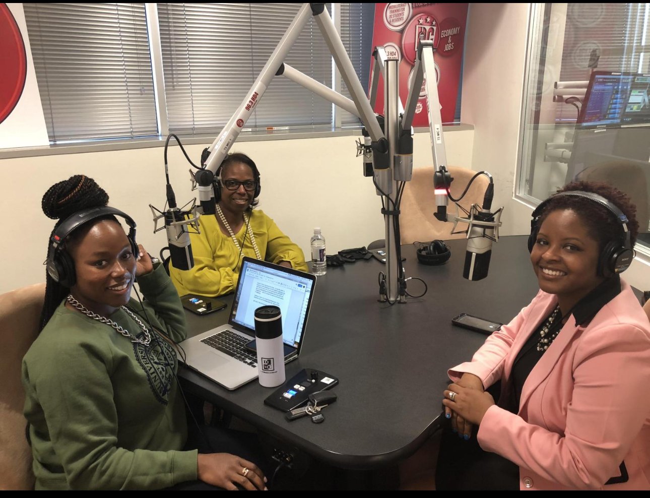 Three women sitting together at a table, equipped with laptops and microphones, focused on their discussion.