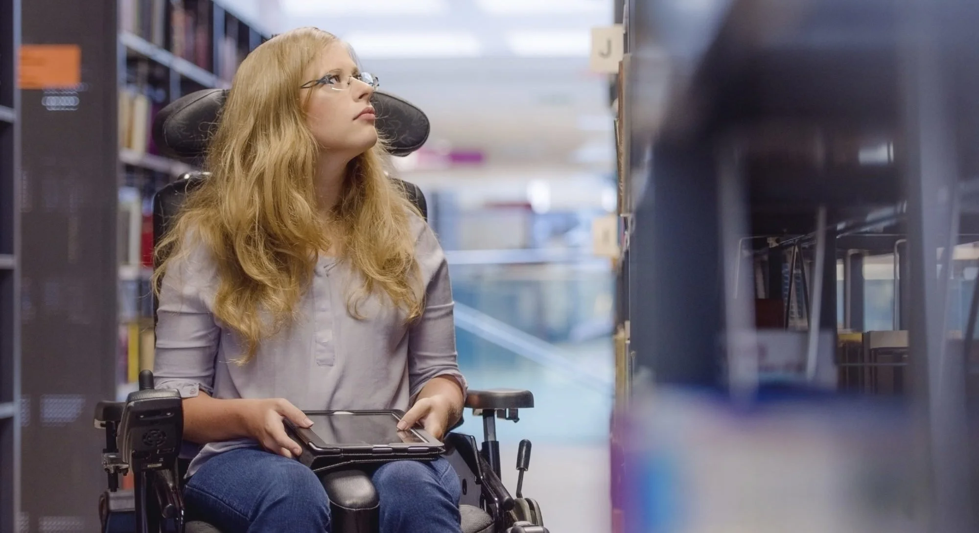 A young woman in a wheelchair is focused on a tablet, engaging with its content thoughtfully.
