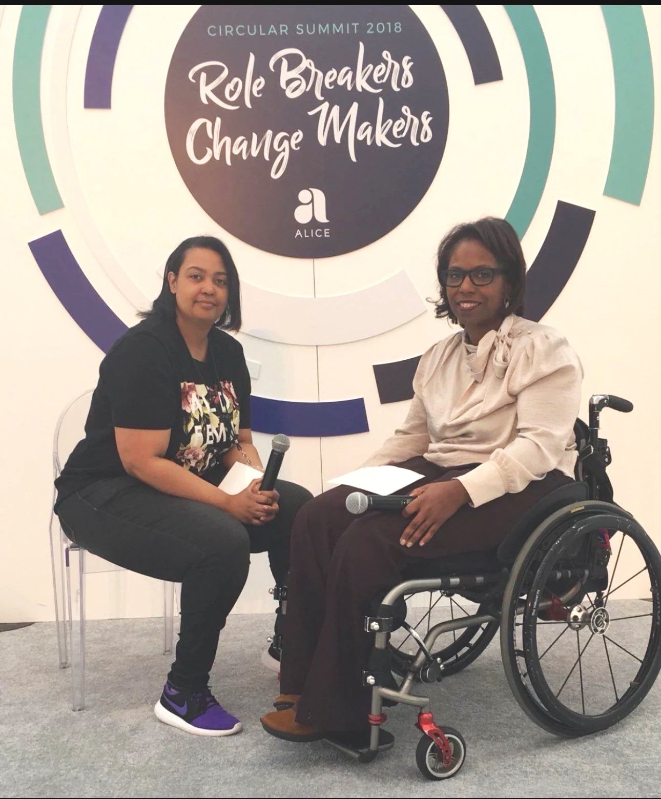 Two women in wheelchairs seated on a stage, engaged in conversation during an event.