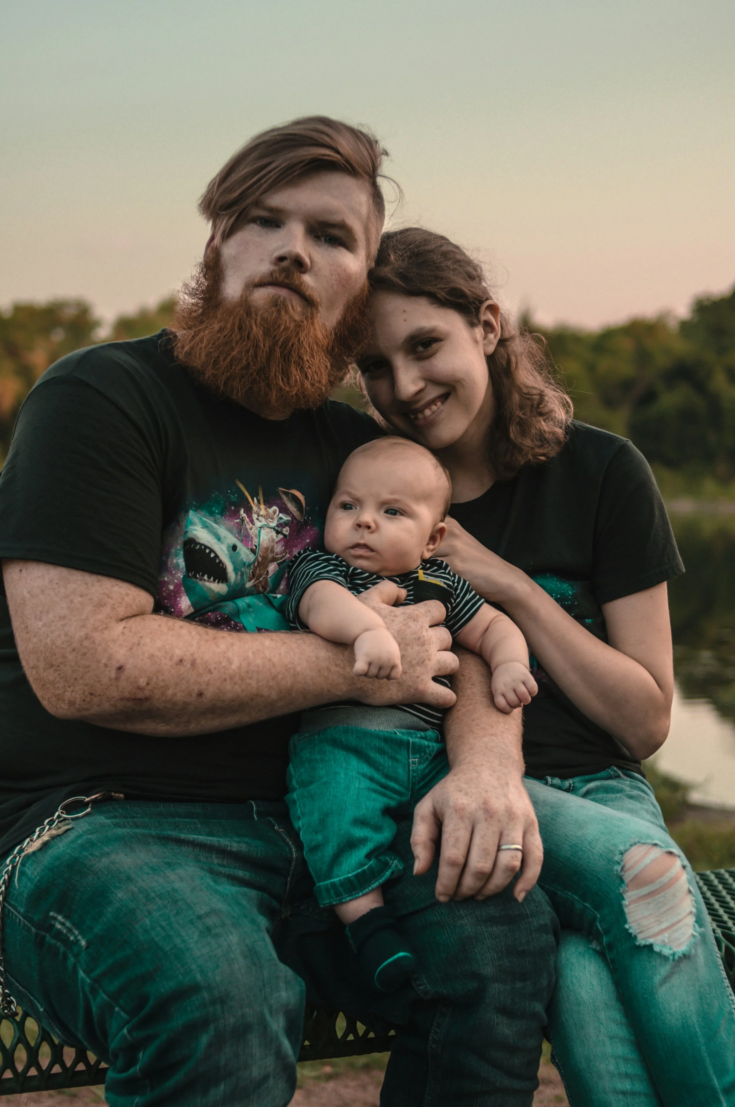 A man and woman stand by a lake, holding a baby, surrounded by nature's beauty.