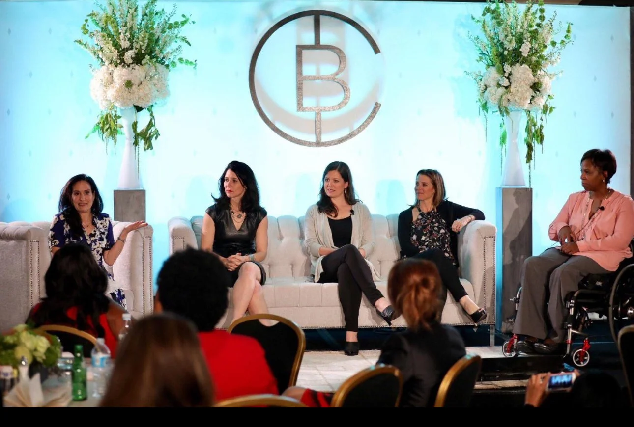 A group of women seated on a stage during a conference, engaged in discussion and sharing insights with the audience.