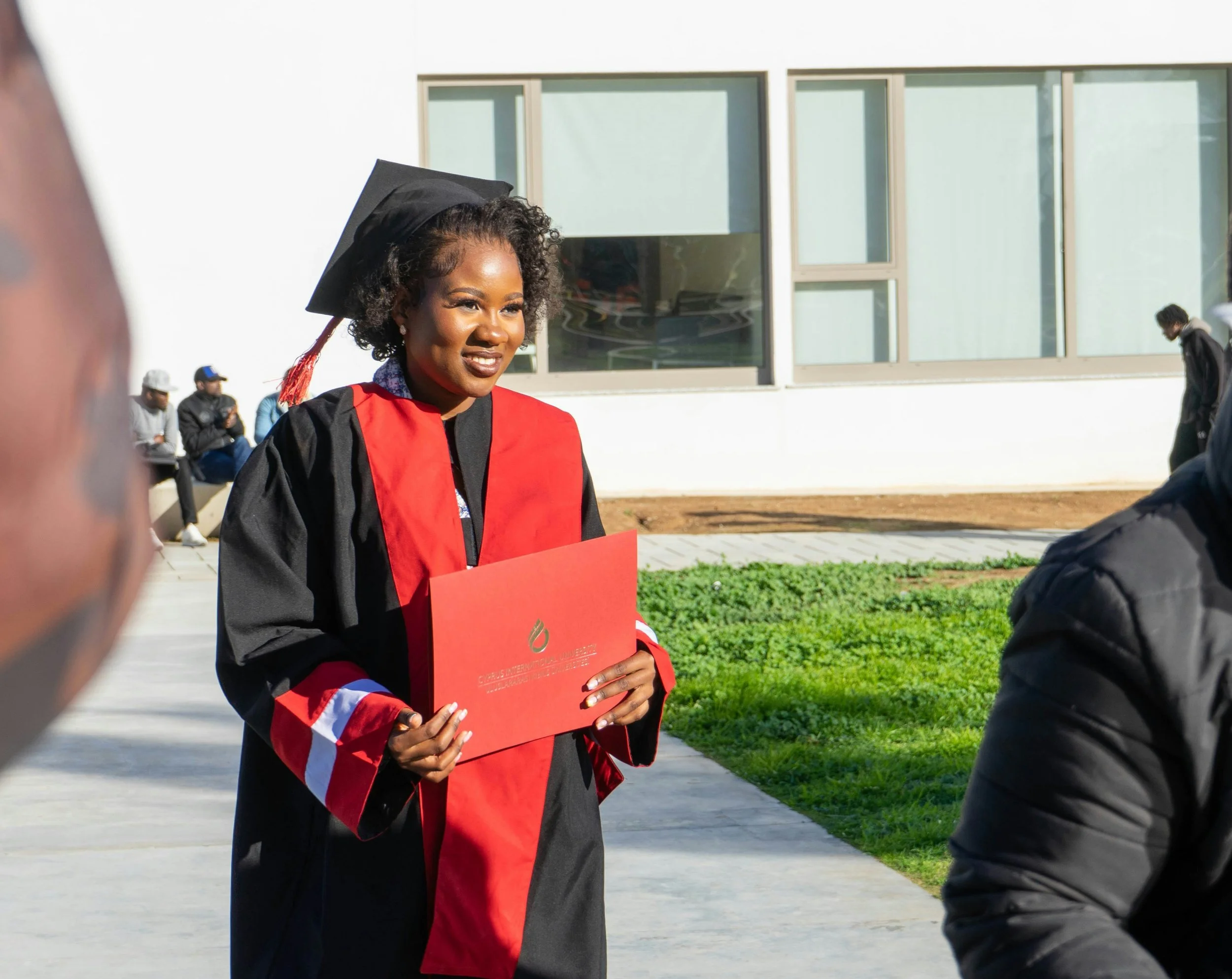 A woman in a graduation gown and red cap proudly holds a red book, celebrating her academic achievement.