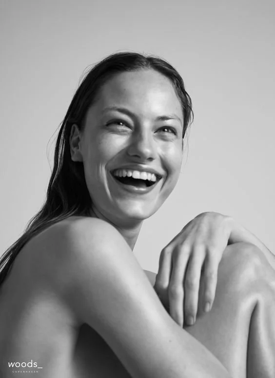Black and white photo of a smiling woman with long hair, sitting with one arm around her knee, looking joyful.