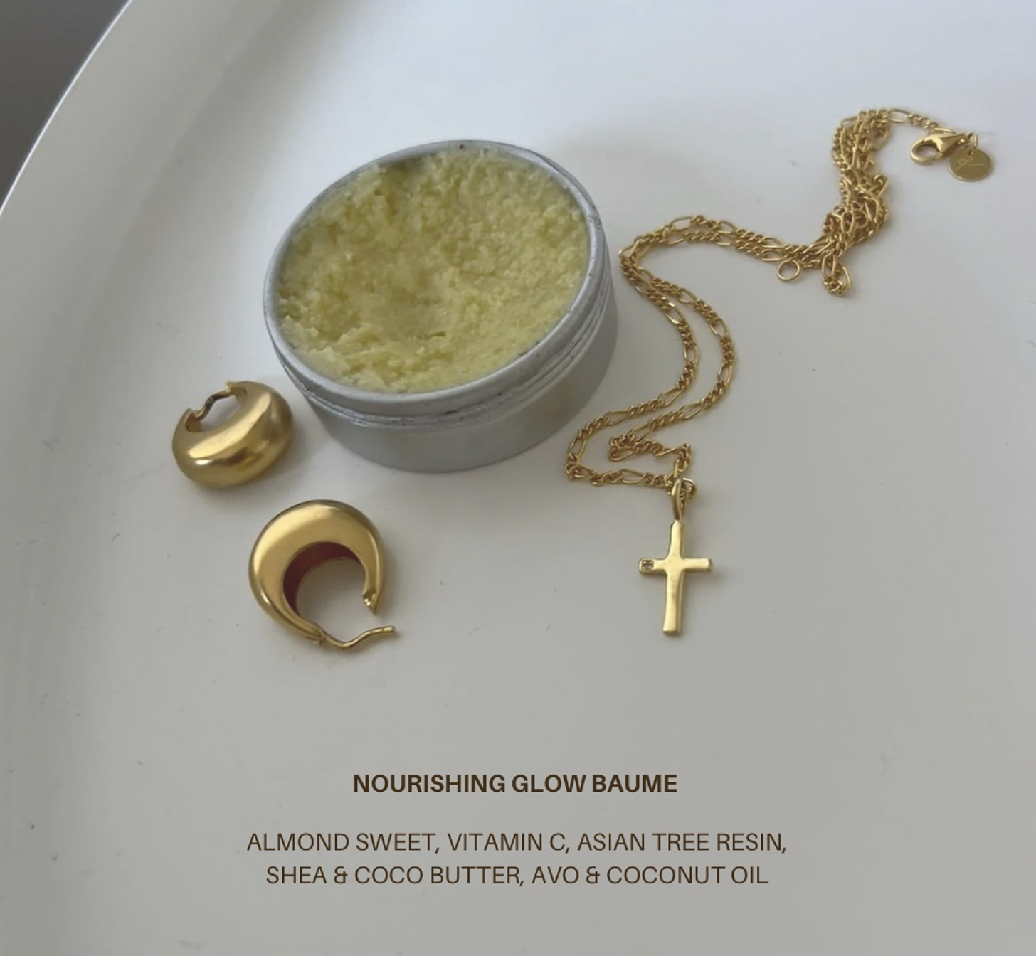 A white tray holding a small jar of yellow nourishing glow balm, gold jewelry including hoop earrings, a chain with a cross pendant, and an advertisement text for a skincare product with ingredients such as almond, vitamin C, Asian tree resin, shea and coconut butter, avocado, and coconut oil.
