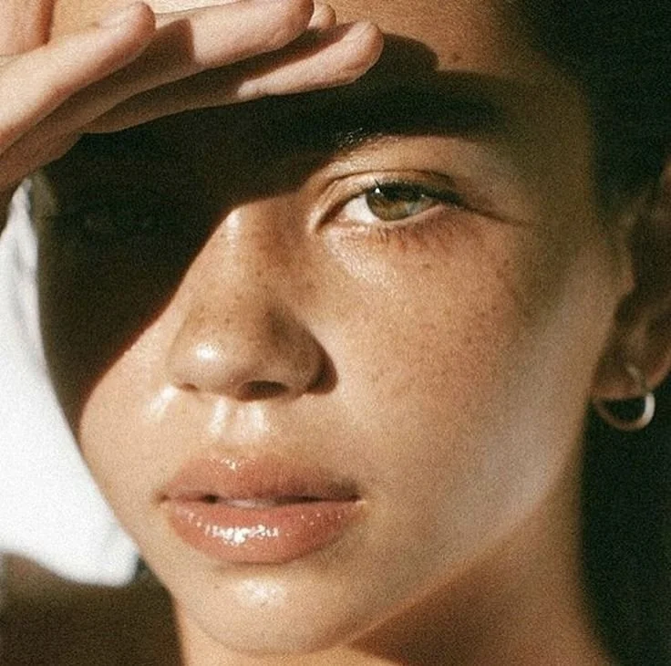 Close-up of a woman with tan skin, freckles, and shiny lips, shielding her eyes with her hand