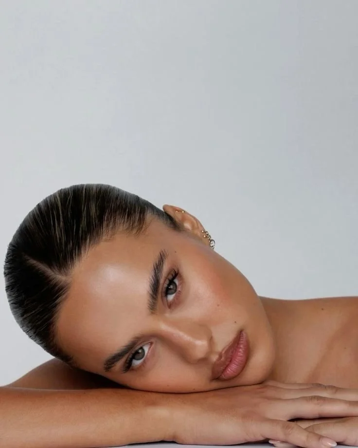 A woman with slicked-back hair resting her face on her arm, looking into the camera with neutral expression, against a plain background.