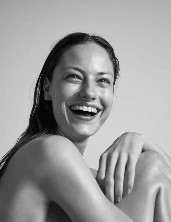 Black and white photo of a woman smiling and laughing, with her hand resting on her knee, looking carefree and joyful.