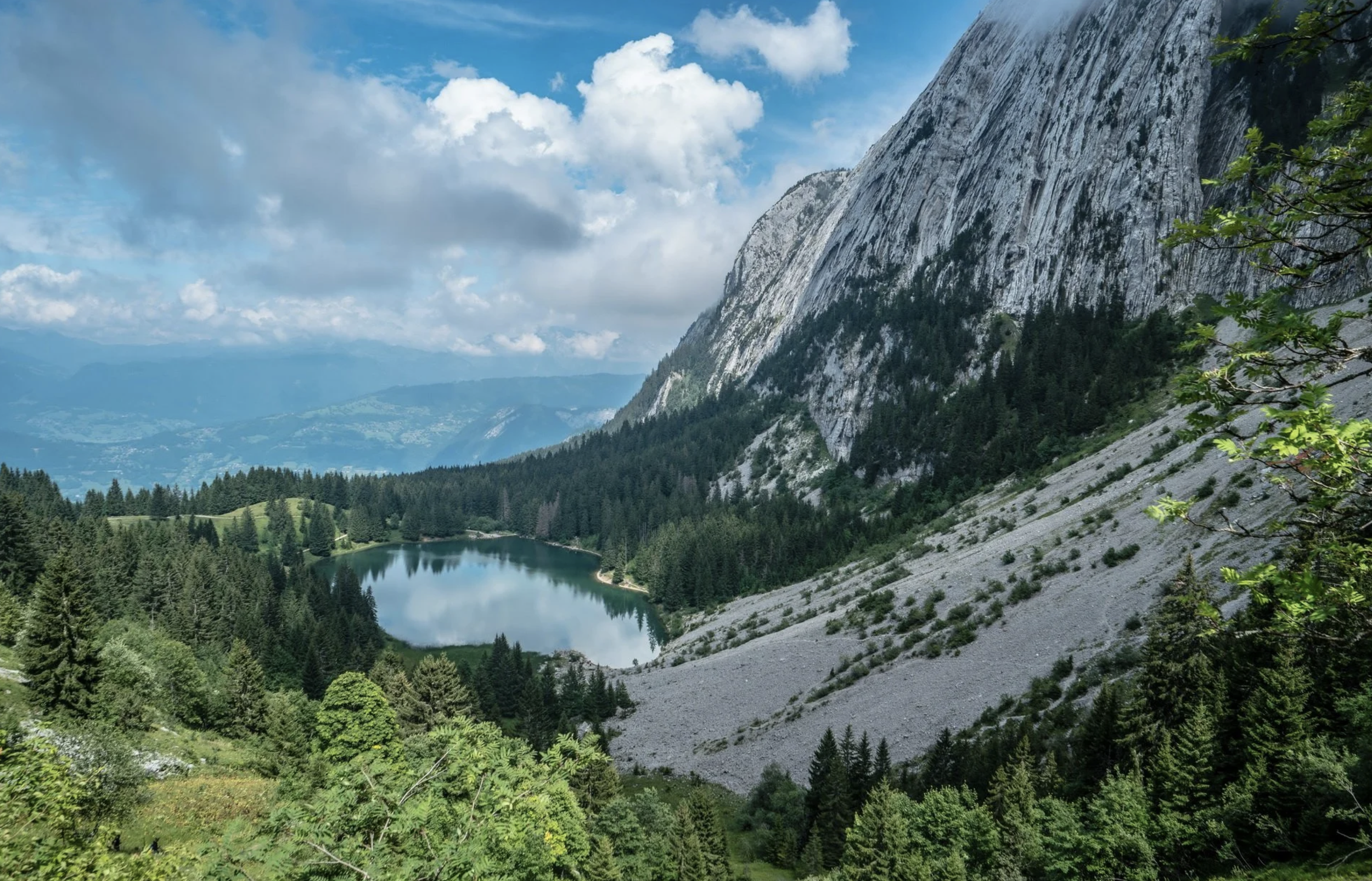 Stunning alpine landscape featuring a crystal-clear mountain lake surrounded by dense pine forests and dramatic limestone cliffs, under a bright blue sky. An ideal natural location for outdoor filming, photography, hiking, and nature travel content i
