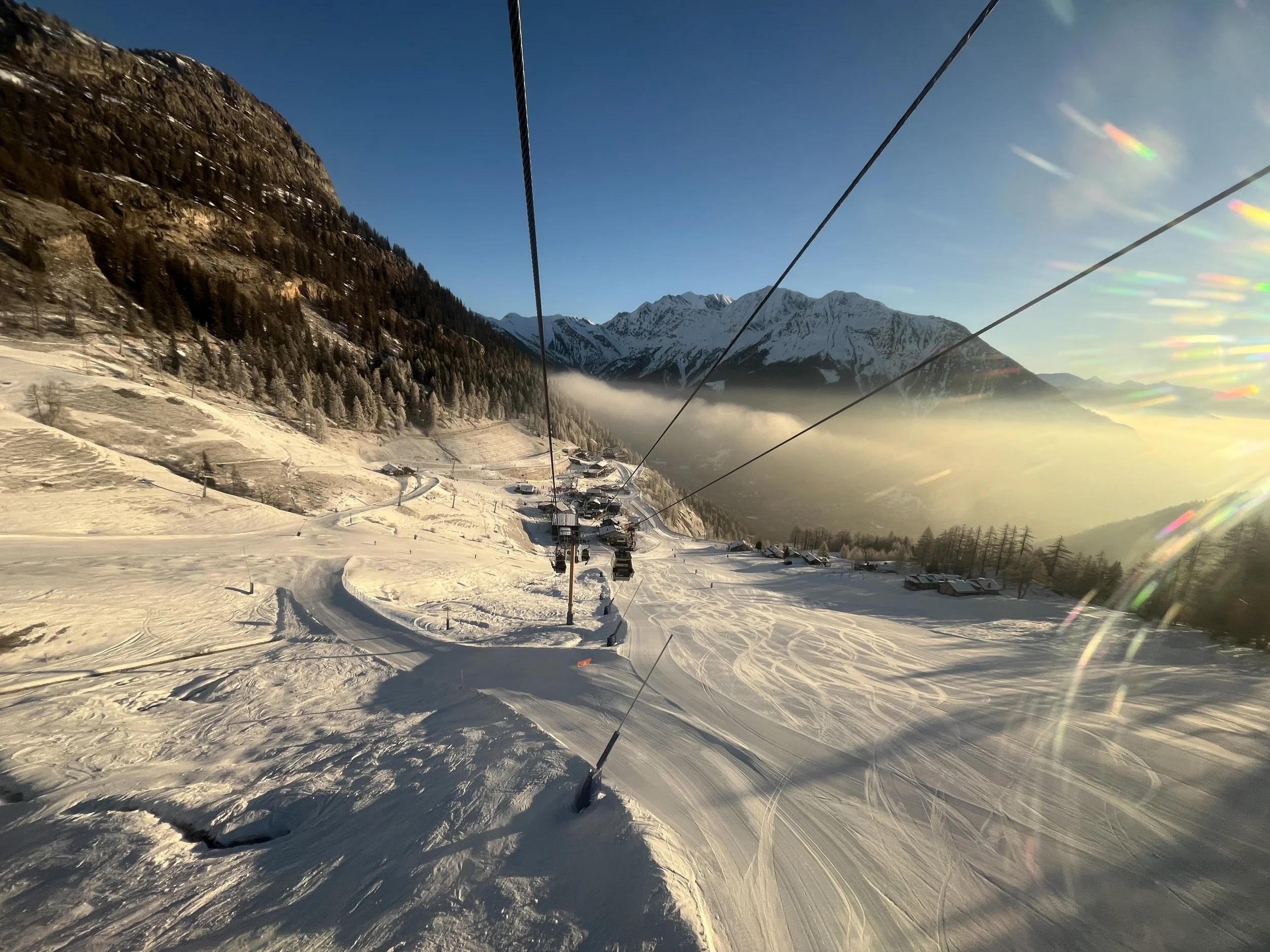 Panoramic winter landscape captured from a ski lift in the Alps, featuring pristine snow-covered slopes, alpine chalets, and dramatic mountain peaks at golden hour. The image showcases a peaceful ski resort scene with fresh ski tracks, frosted trees,