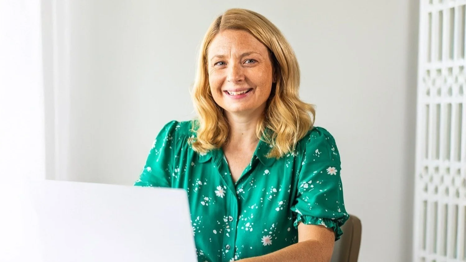 A woman with blonde wearing a green blouse with white floral patterns, smiling while sitting at a desk with a laptop.