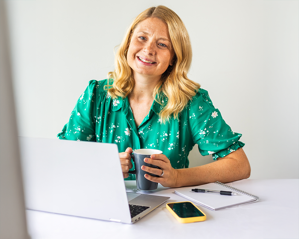 A woman with blonde hair, wearing a green floral blouse, sitting at a desk with a laptop, a notepad, a pen, a smartphone, and holding a coffee mug, smiling