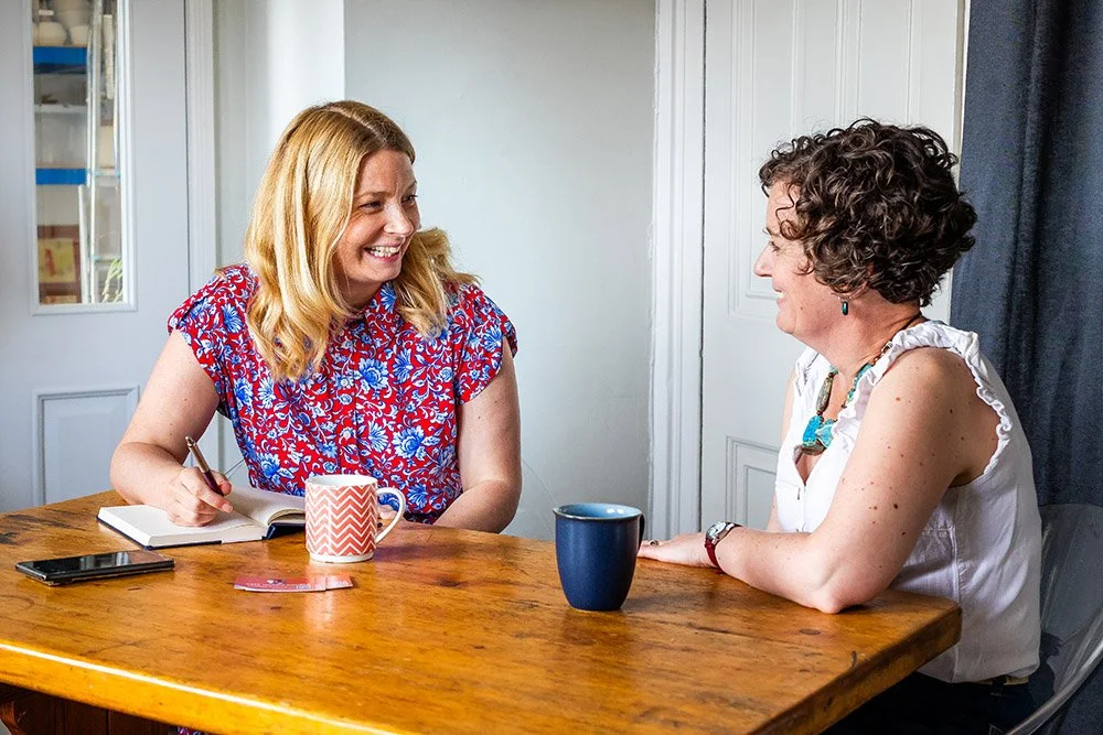 Two women are sitting at a wooden table, smiling and talking. One has blonde hair and is wearing a red and blue floral shirt, taking notes with a pen and notebook. The other has curly hair and is wearing a white sleeveless top with a turquoise necklace, and has a blue mug in front of her. There is also a black smartphone on the table.