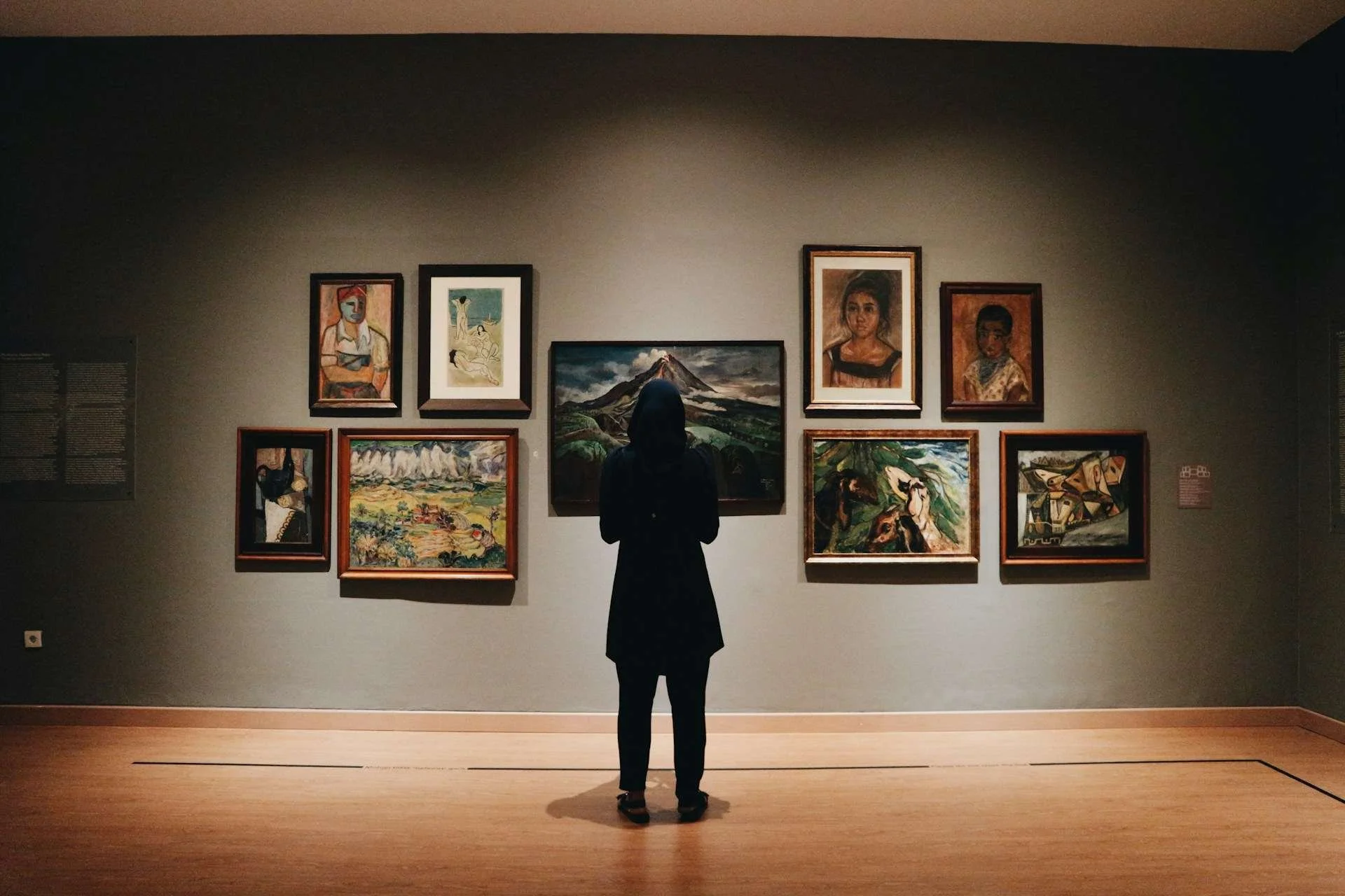 We see a woman in black pants, black coat, and black headscarf from behind. She is standing in front of a wall of paintings in a gallery of an art museum. We cannot see her face, so we do not know which oil painting has captured her attention.