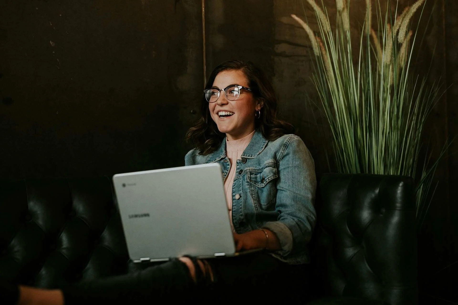 Employer brand is now consumed in conversations with AI chatbots. Photo: young woman with turtleneck sweater and denim jacket working on a laptop. She's seated on a leather couch and smiling at someone out of frame.