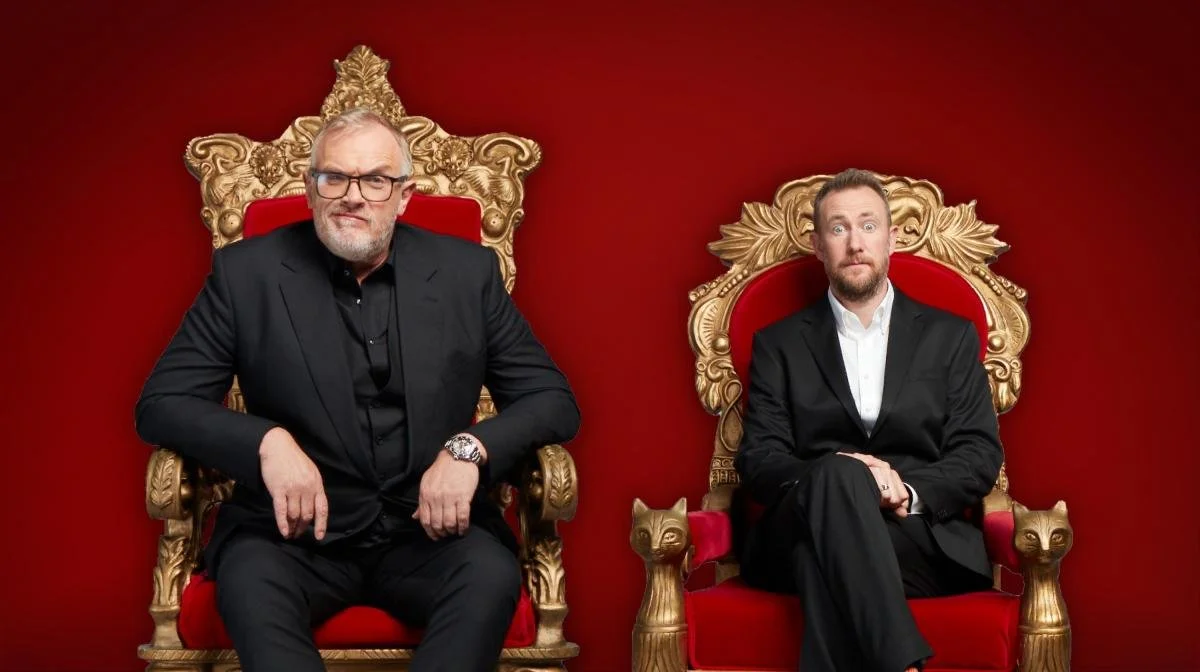 Publicity photo for the British TV Show Taskmaster. Host Greg Davies and Assistant Alex Horne are both wearing black suits, sitting in red and gold thrones, and seated against a deep red backdrop. Greg Davis wears eyeglasses, a large watch, and black