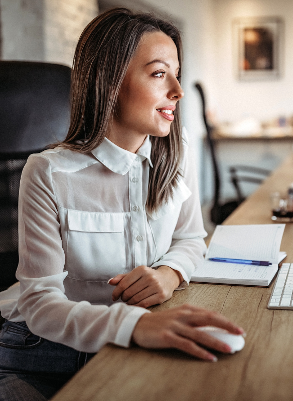A young woman with shoulder-length brown hair, wearing a white blouse, sitting at a wooden desk with a computer mouse, notebook, and pen, in a modern office setting.