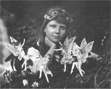 A black and white composite photograph from 1917. A girl wearing a flower crown looks wistfully into the camera. In the foreground, a trick using paper cutouts makes it look like fairies are dancing around the girl.