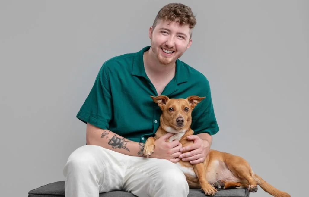 A young white man in his 20s with a light red-brown beard and curly hair sits in front of a grey background. We wears white pants and a dark emerald shirt. A small mixed-breed tan-colored dog with pointy ears and a white chest sits on his lap