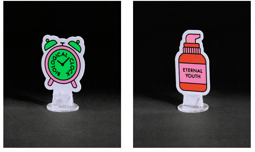 Two pieces from the Billie "No Worries If Not!" Board Game. One is a token of an alarm clock that says "biological clock" and one is a bottle of serum that says "eternal youth."