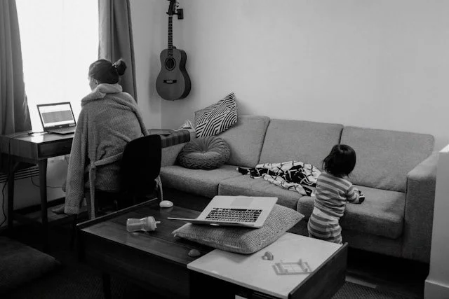 Black and white photo of a small living room. Against the window is a small desk with a laptop computer. A young woman is working at the desk. Behind her, a toddler is playing alone, leaning against a couch. On the coffee table is a baby bottle and a