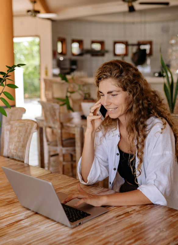 A white woman in her 30s or 40s, has natural eyebrows and curly red brown hair down to her waist. She sits in the living area of a home in neutral hues and talks on her mobile phone while looking at her laptop. She looks confident and happy.
