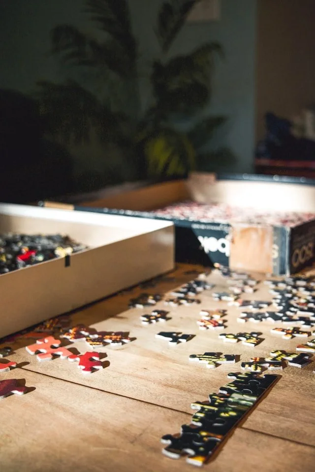 A cozy domestic scene shows a secondhand jigsaw puzzle in progress, the pieces are spread across a light wooden table. Two halves of the puzzle box rest nearby, one has a piece of packing tape to mend a tear. The shallow depth of field creates a soft