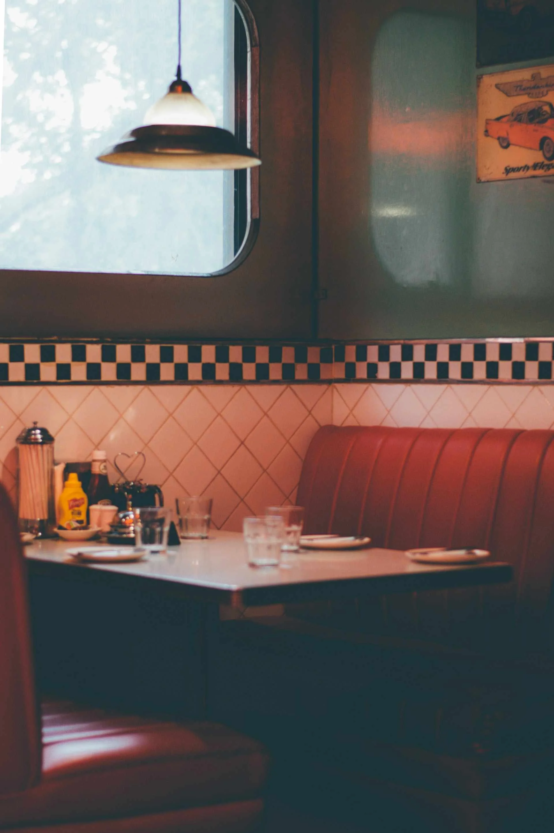 Photograph of a classic American diner booth. There are red pleather banquette seats, black and white checked tile border, and rounded, chrome-lined windows. The table top includes a canister of paper straws and condiments.