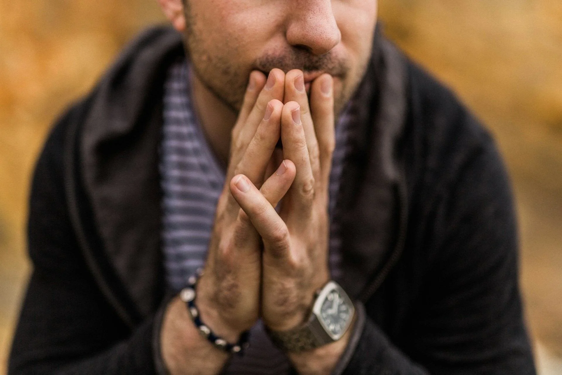 We see the lower half of a man's face and his midsection as he sits pensively. He wears a striped shirt and a hoodie. His hands are clasped in front of him with this fingers covering his mouth and chin. He wears a bracelet on one wrist and a watch on