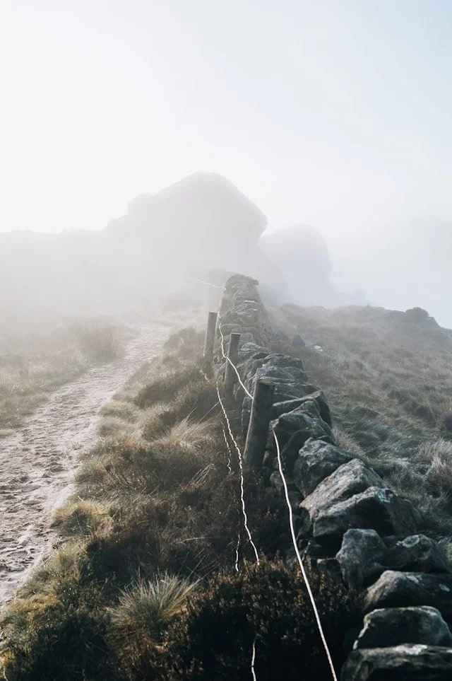 Photography of a foggy trail with rocks silhoutetted in the distance. The trail looks muddy. A simple wire fence runs along the old stone wall. Photo taken in Skipton, UK