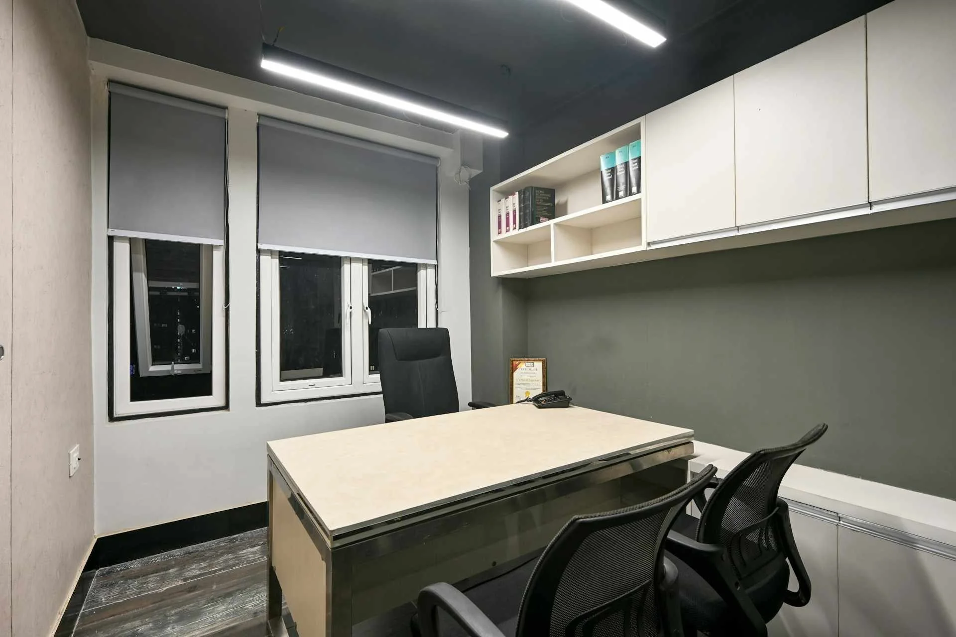 Empty corporate office at night time. There is a desk with one office chair on one side, and two chairs across the desk. The only thing on the desk is a telephone. The room is lit by harsh fluroscent lighting.