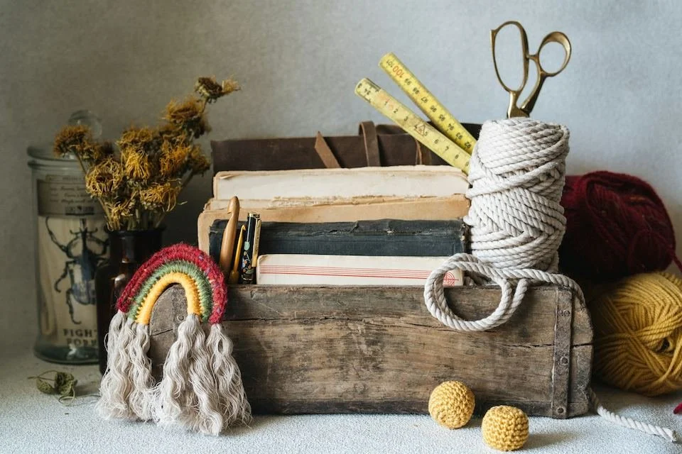 An assortment of analog hobby supplies are organized in a weathered wooden tray. There is a leatherbound journal, crochet hooks, measuring sticks, large gold shears and macrame cord in the slots. A hand-macramed rainbow hangs off one corner and the e