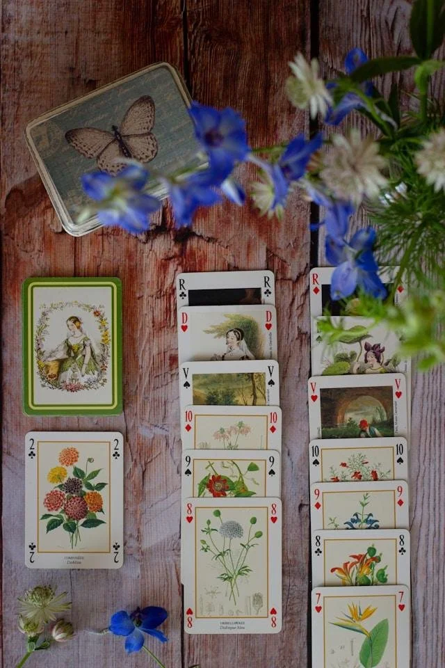 A dreamy arrangement of vintage botanical played cards arranged in a solitaire tableau agsinst weathered wooden planks. Fresh blue and white flowers appear unfocussed in the top right corner of the frame.