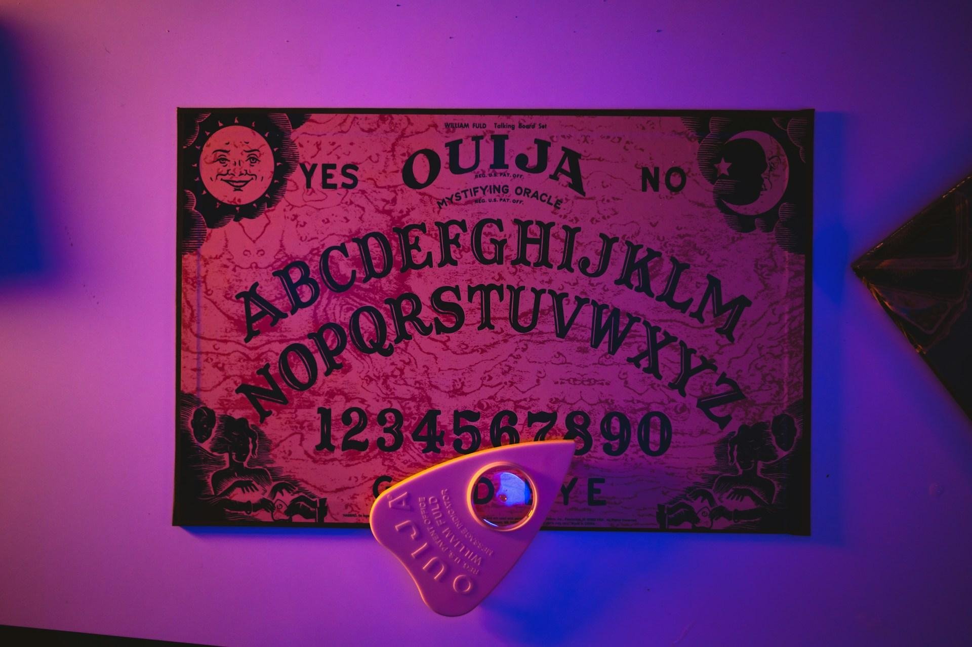 A classic Ouija Board and planchette are set up on a table in eerie purple and blue lighting. The top of the Ouija Board reads "William Fuld Talking Board Set. Mystifying Oracle."