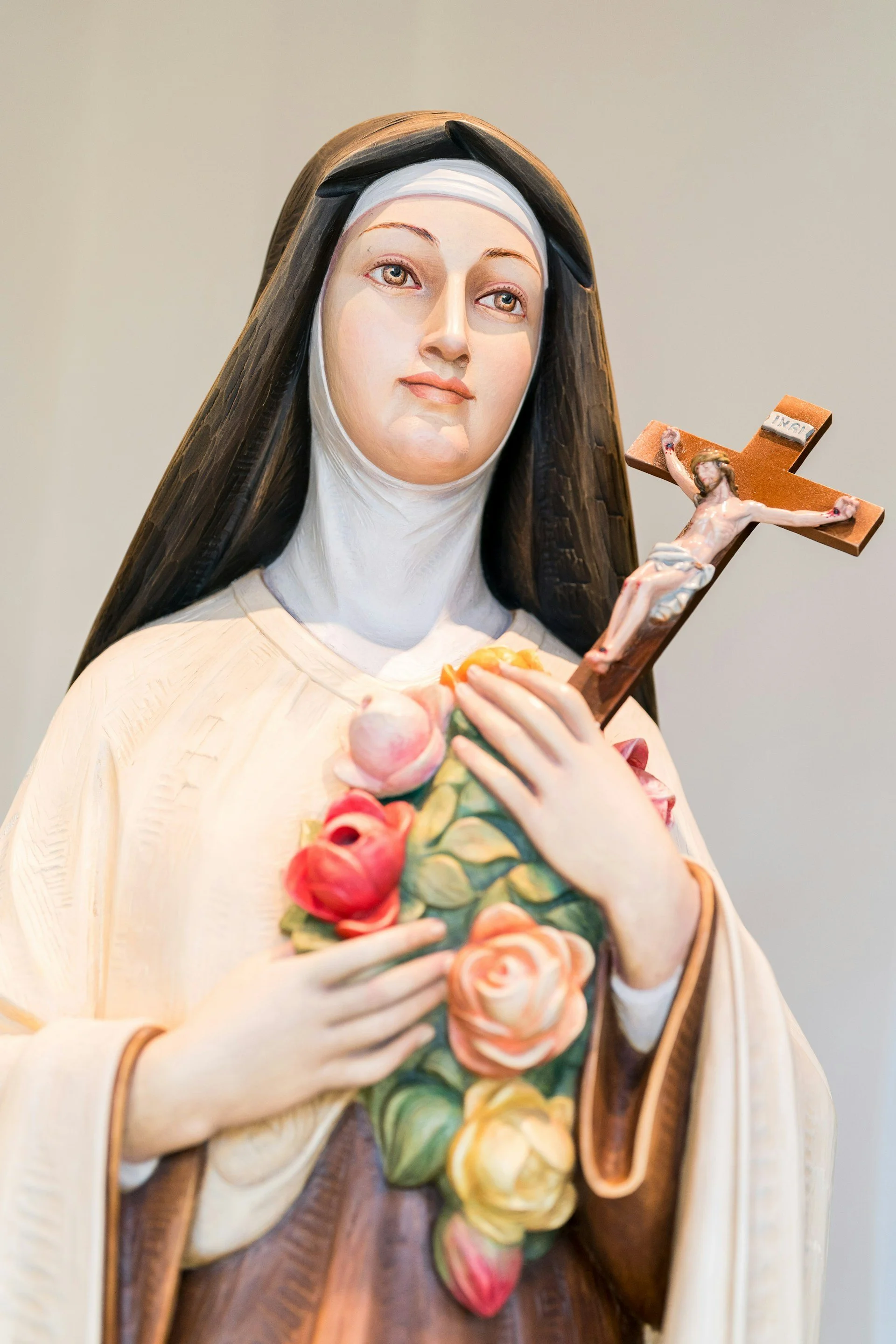 Photograph of a statue of Saint Therese of Lisieux, taken at St. Therese of Lisieux Roman Catholic Church in Montauk, NY. She wears a white robe and a black habit. She's holding a crucifix covered in a spray of colorful roses.