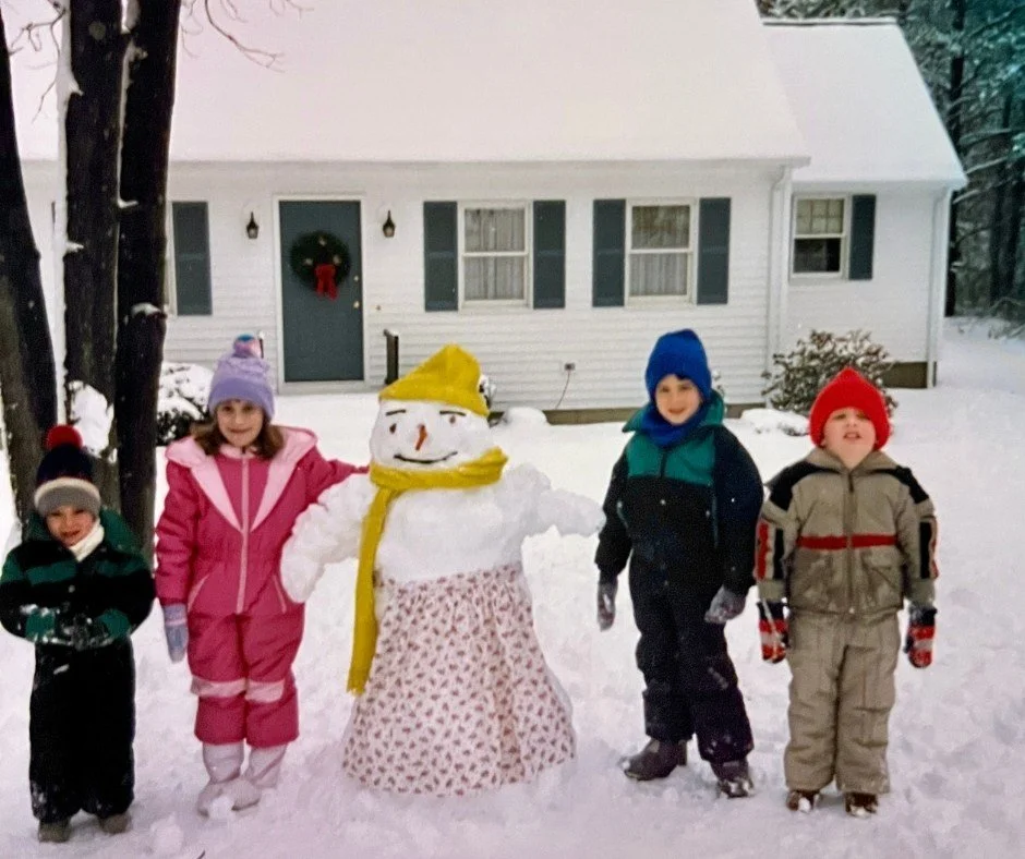 Photo from the late 1980s of kids posing with a snowman in front of a Cape Cod style house in New England. Four kids wear snowsuits, hats, scarves and gloves. The snowwoman wears a hat, scarf, and apron with a carrot nose.