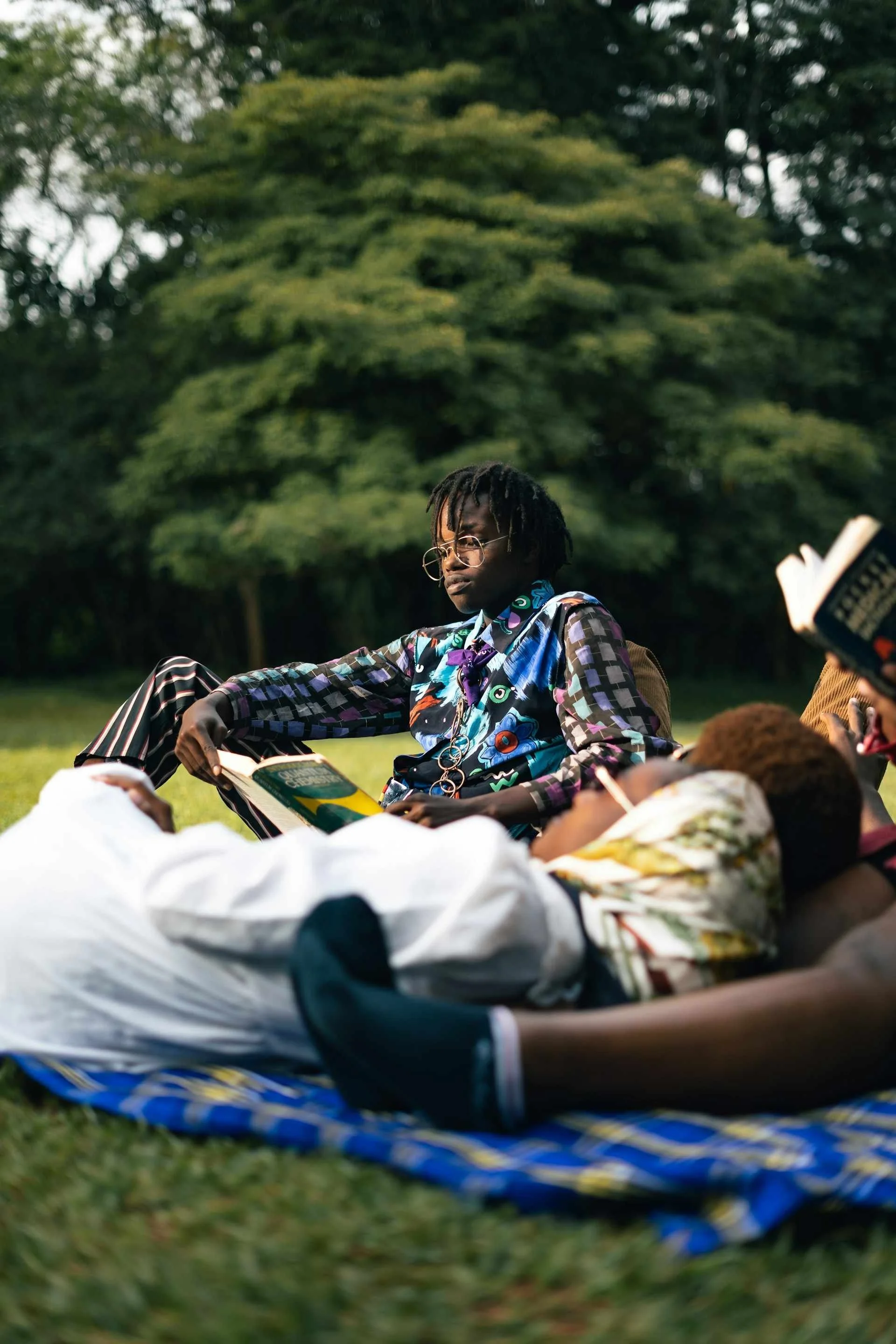 Some Black young adults are lounging on a picnic blanket in a park. Two of the people are reading different books; they're in community together while still doing their own thing. It looks like a warm, and peaceful way to spend time together.