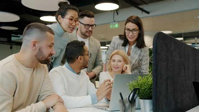 Stage photo of a group of professionals gathered around a single laptop. Six people are gathered and listening to the man in the center of the photo (owner of the laptop). The author asserts no actual work has ever been accomplished like this.