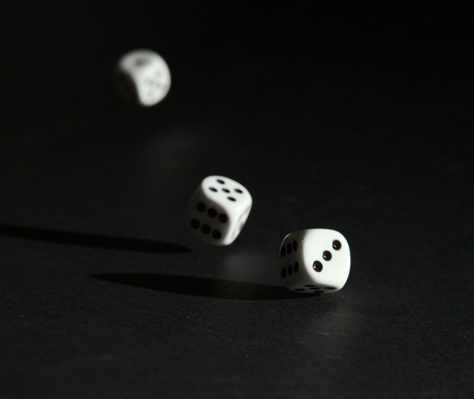 Photo of three white dice mid-throw. The white dice have black pips and are set against a black background. The two dice in the foreground cast shadows, but the third dice is blury and will be the last to land. On the foreground dice we observce pips