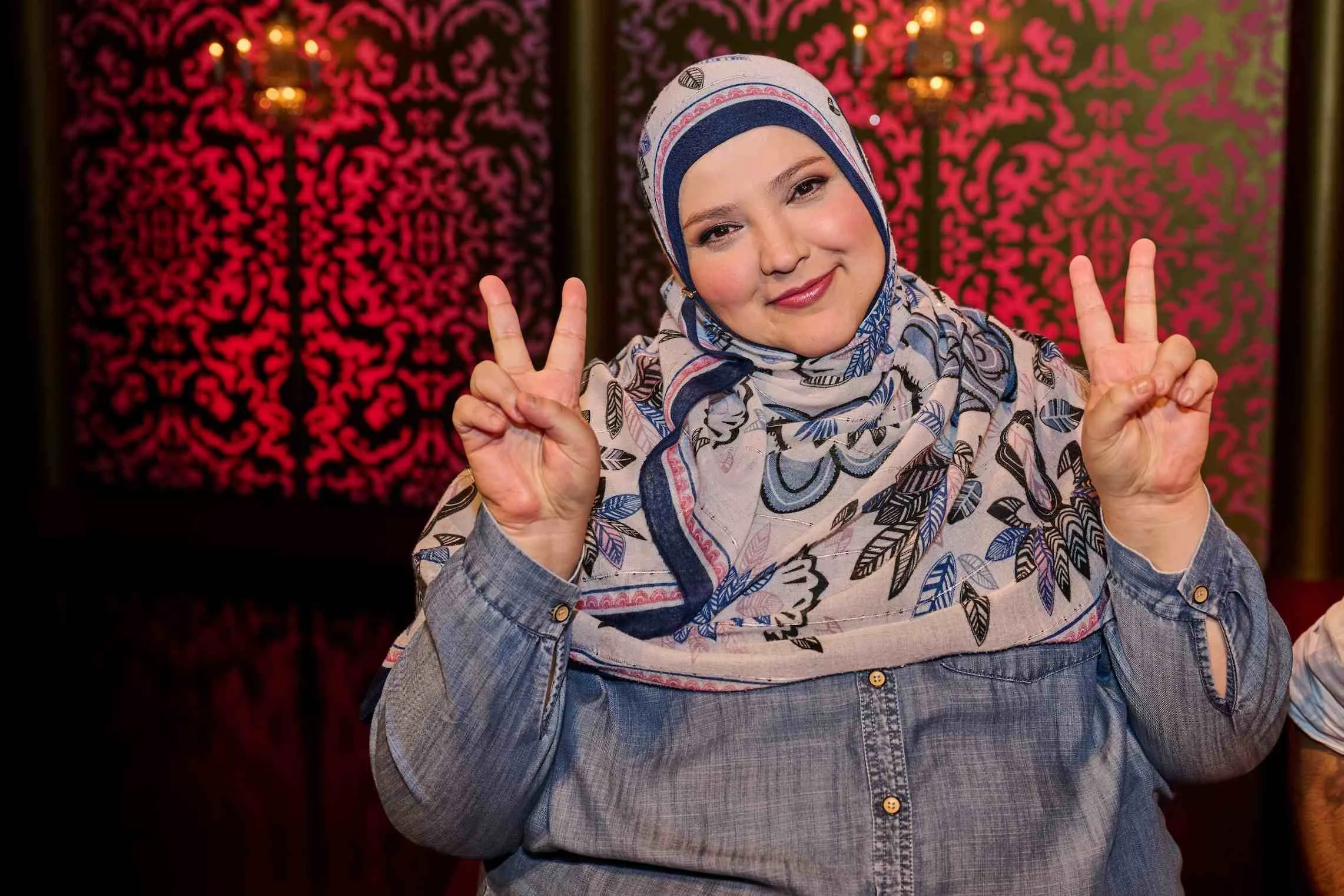 Photo of Taskmaster contestant Fatiha El-Ghorri from Series 19. A middle-aged woman wears a denim shirt and a floral-print hijab. She has rosy cheeks and pink lipstick. Her smile makes her appear cute and cuddly.
