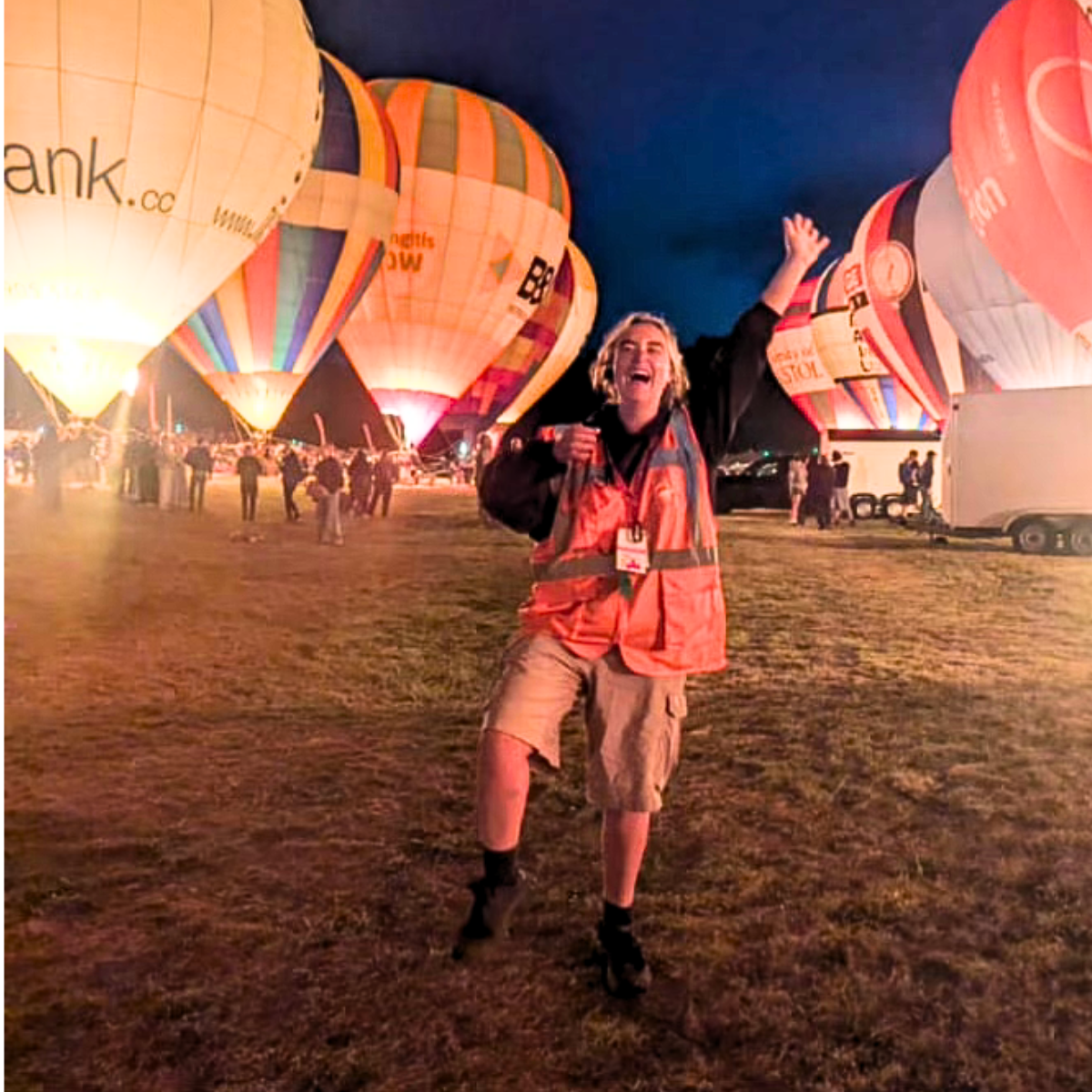 Person wearing a high-visibility vest standing in a field at night with illuminated hot air balloons and a crowd in the background.