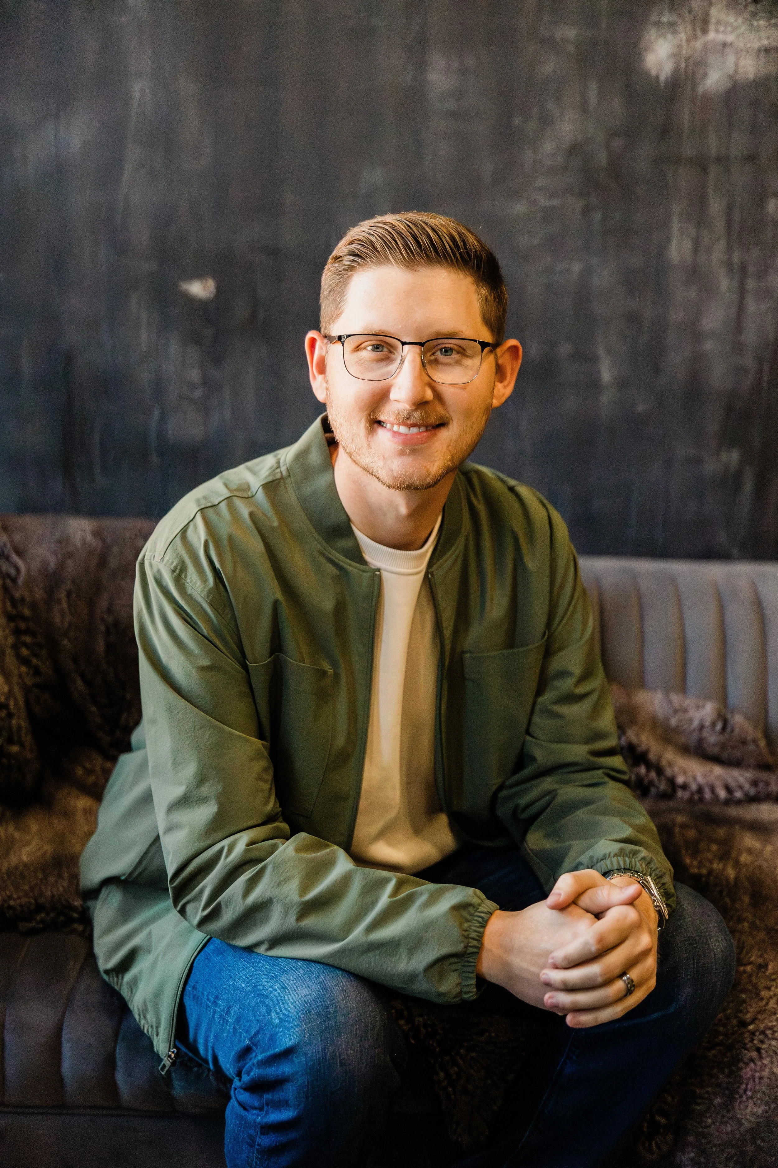 A young man with glasses and light brown hair, wearing a green jacket and a beige shirt, sitting on a couch with a dark textured background, smiling and looking at the camera.