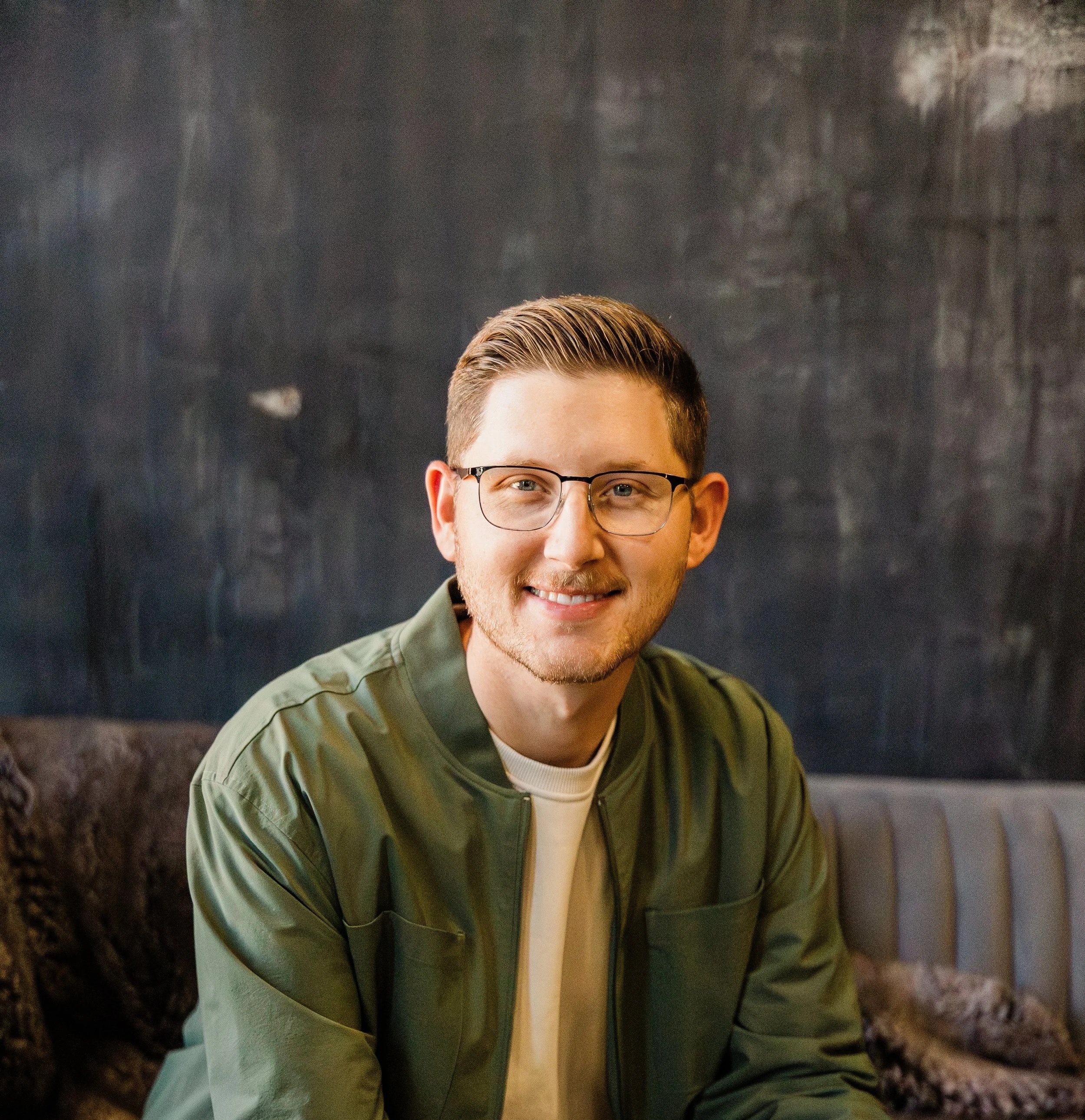 A young man with brown hair, glasses, and a smile, wearing a green jacket and white shirt, sitting against a dark, textured wall.