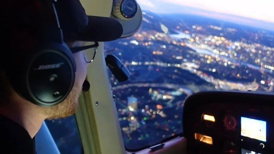 A person flying in a plane with headphones, looking out at a cityscape during sunset.