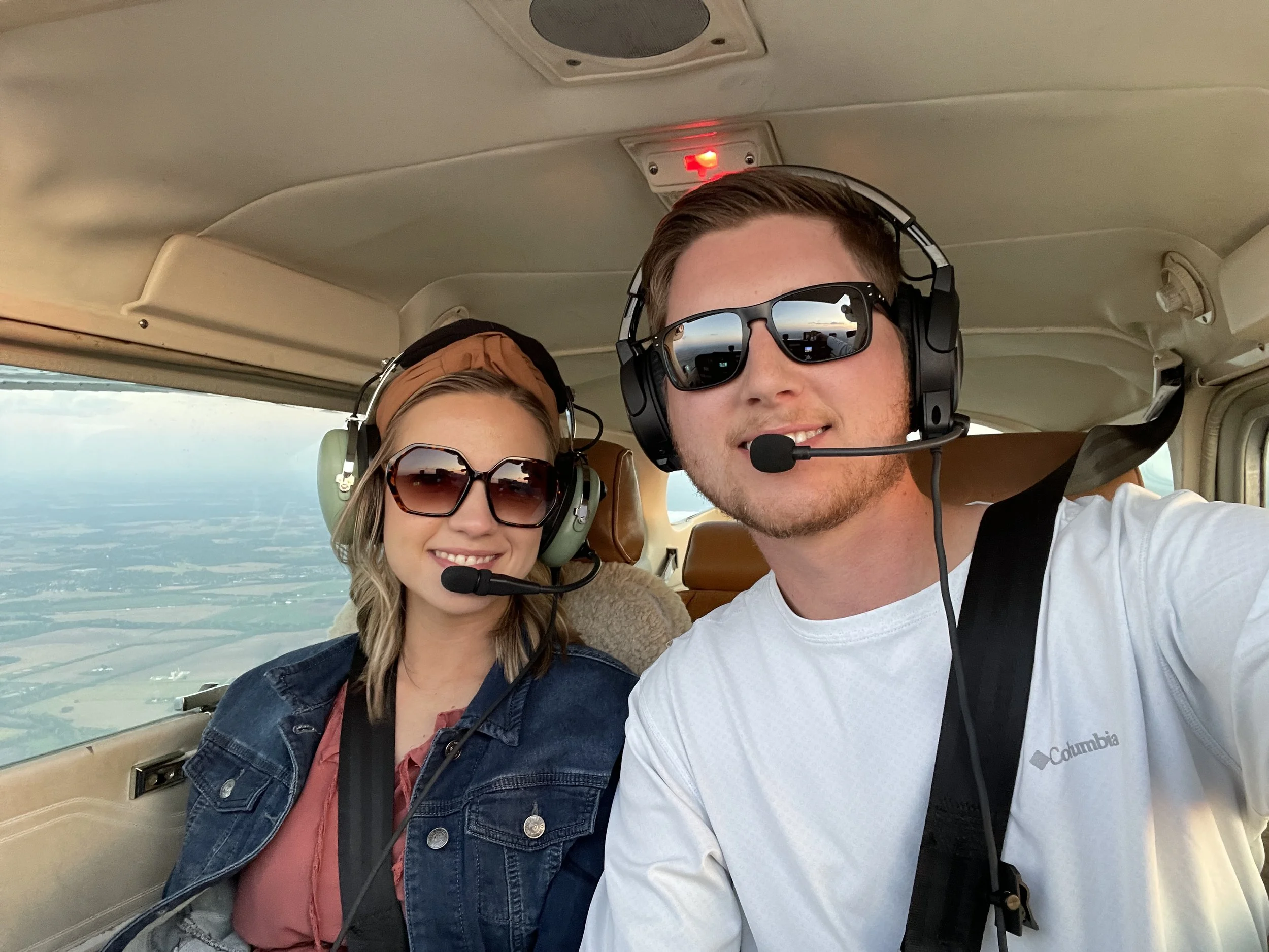Two people, a man and a woman, taking a selfie inside an airplane cockpit. They are wearing headsets with microphones and sunglasses. The man is smiling and has short hair, while the woman has blonde hair and is also smiling. The background shows the