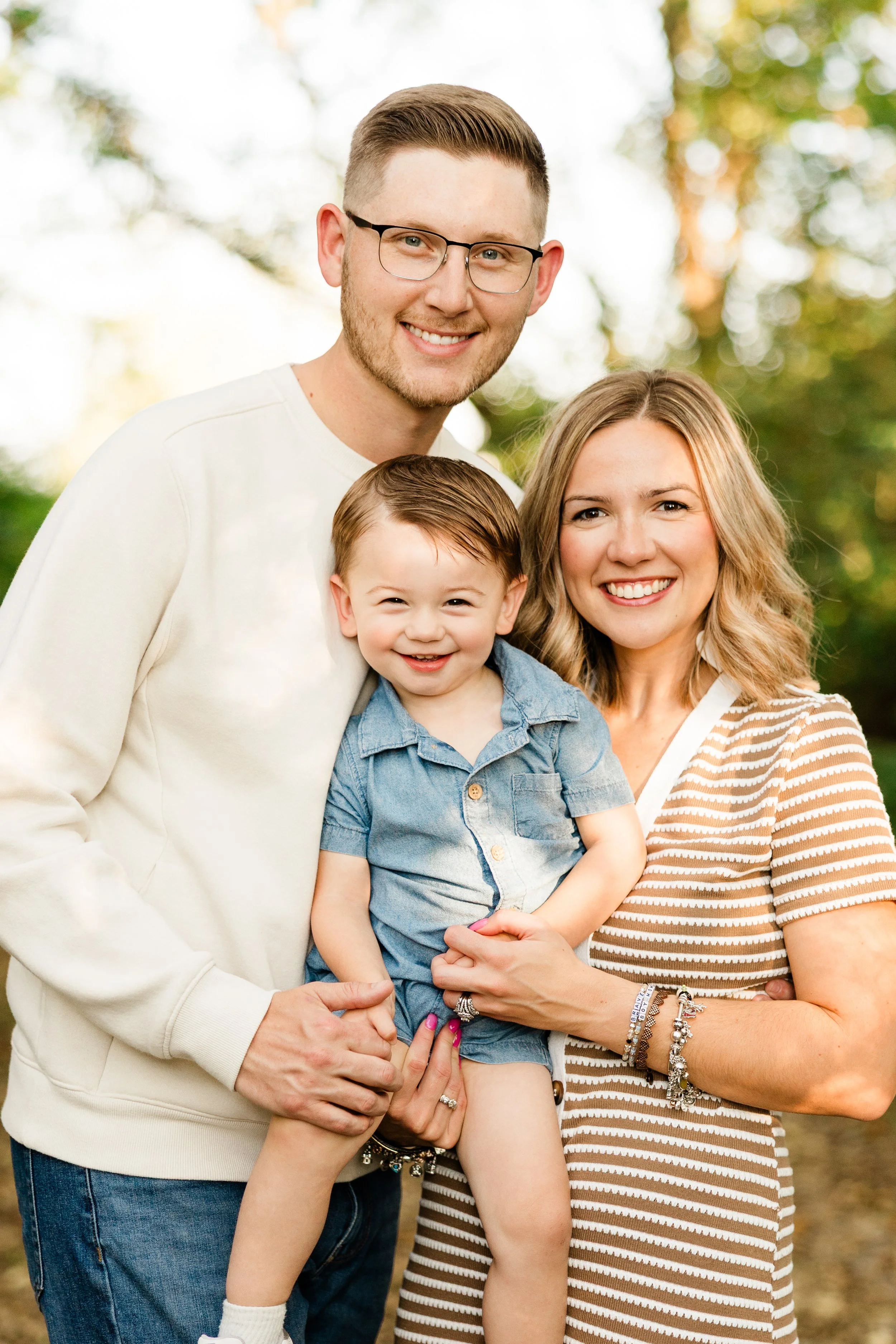 A smiling family of three standing outdoors with trees in the background. The man has glasses, light brown hair, and a beard. The woman has long, wavy blonde hair, and is wearing a striped top. The child, a young boy, has short light brown hair and i