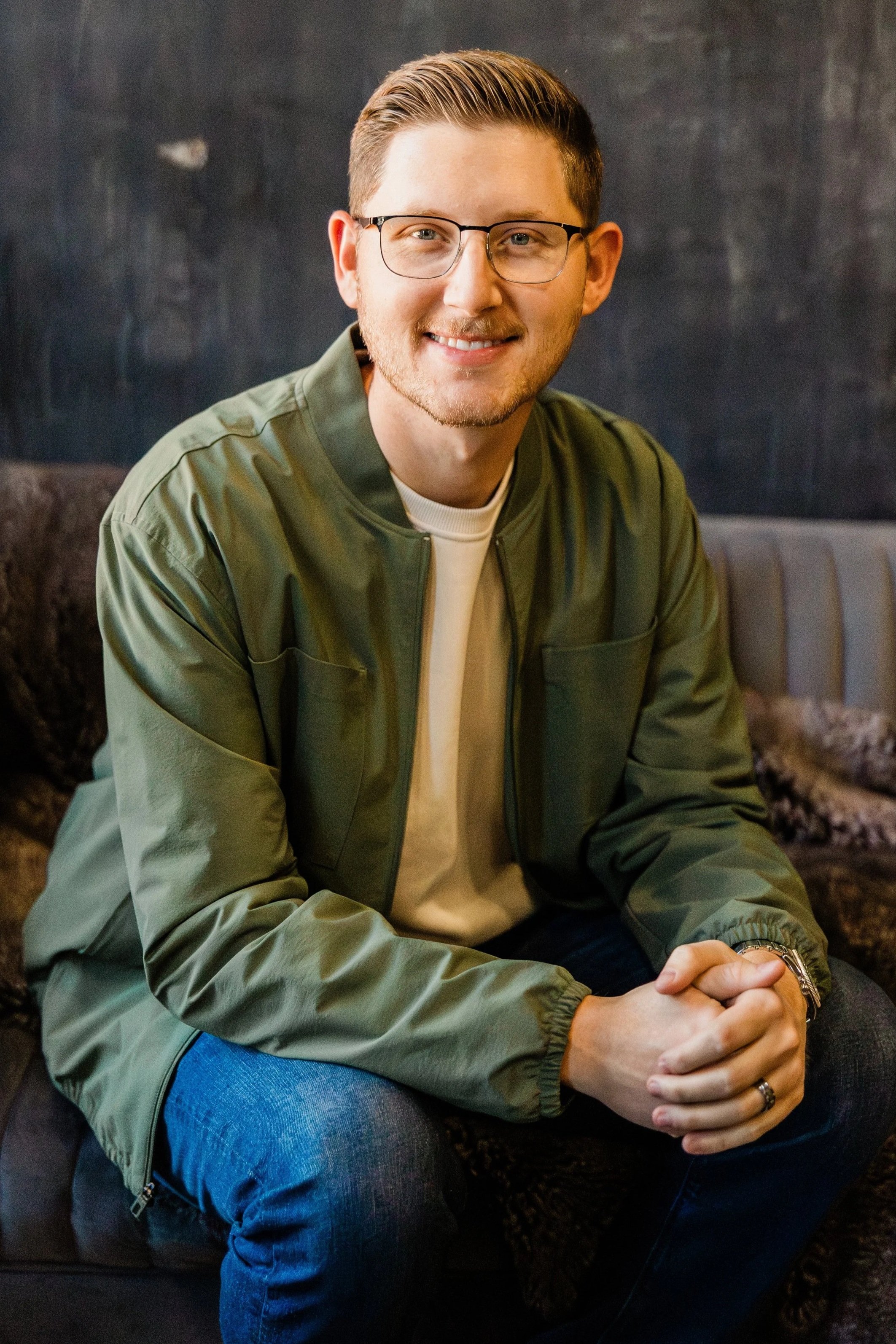 A man with glasses, light brown hair, and a short beard smiling while seated indoors on a dark textured surface, wearing a green jacket and blue jeans.