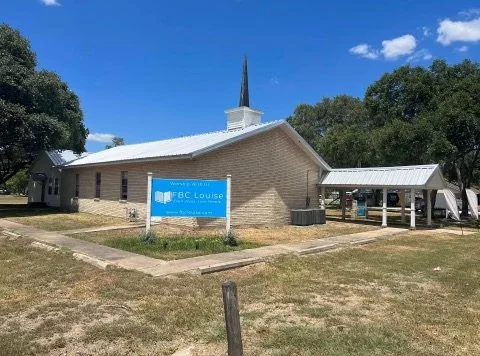 A small brick church with a steeple and a blue signboard in front that reads 'BC Louise', surrounded by trees under a partly cloudy sky.