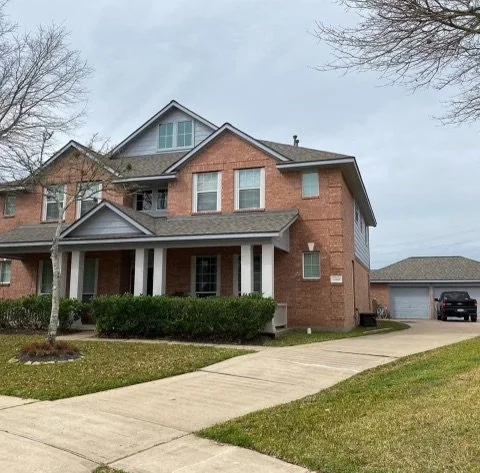 A two-story brick house with a front porch, surrounded by a well-maintained lawn and a paved driveway leading to a garage in a suburban neighborhood.