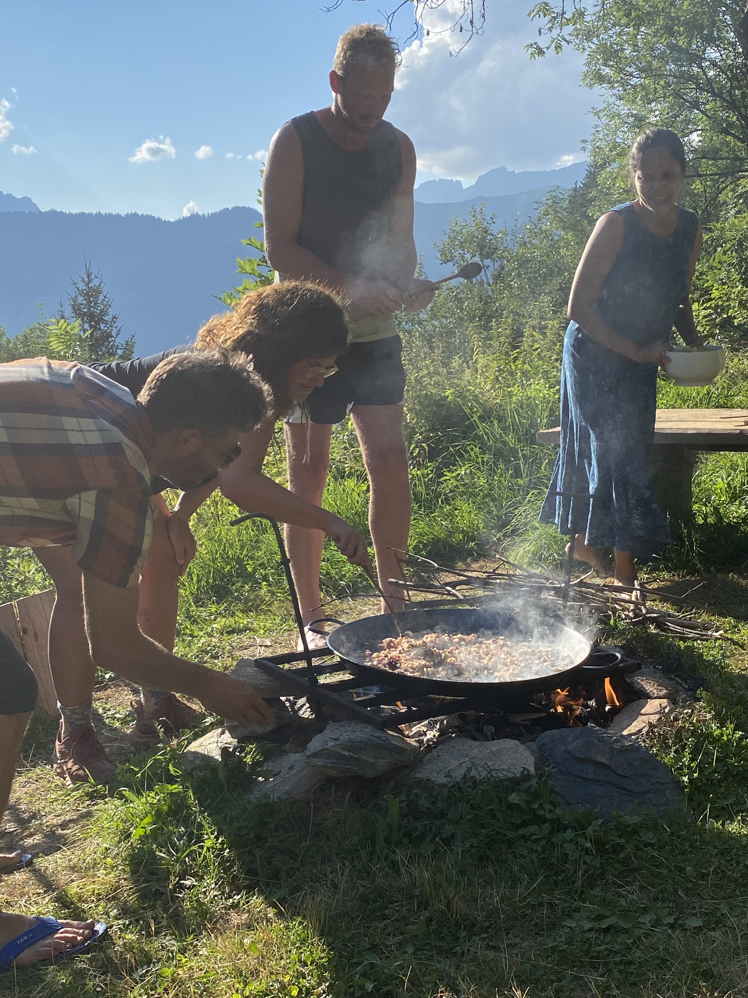 Four people outdoor around a campfire in a wooded mountain area, cooking and preparing food, with mountains and blue sky in the background.
