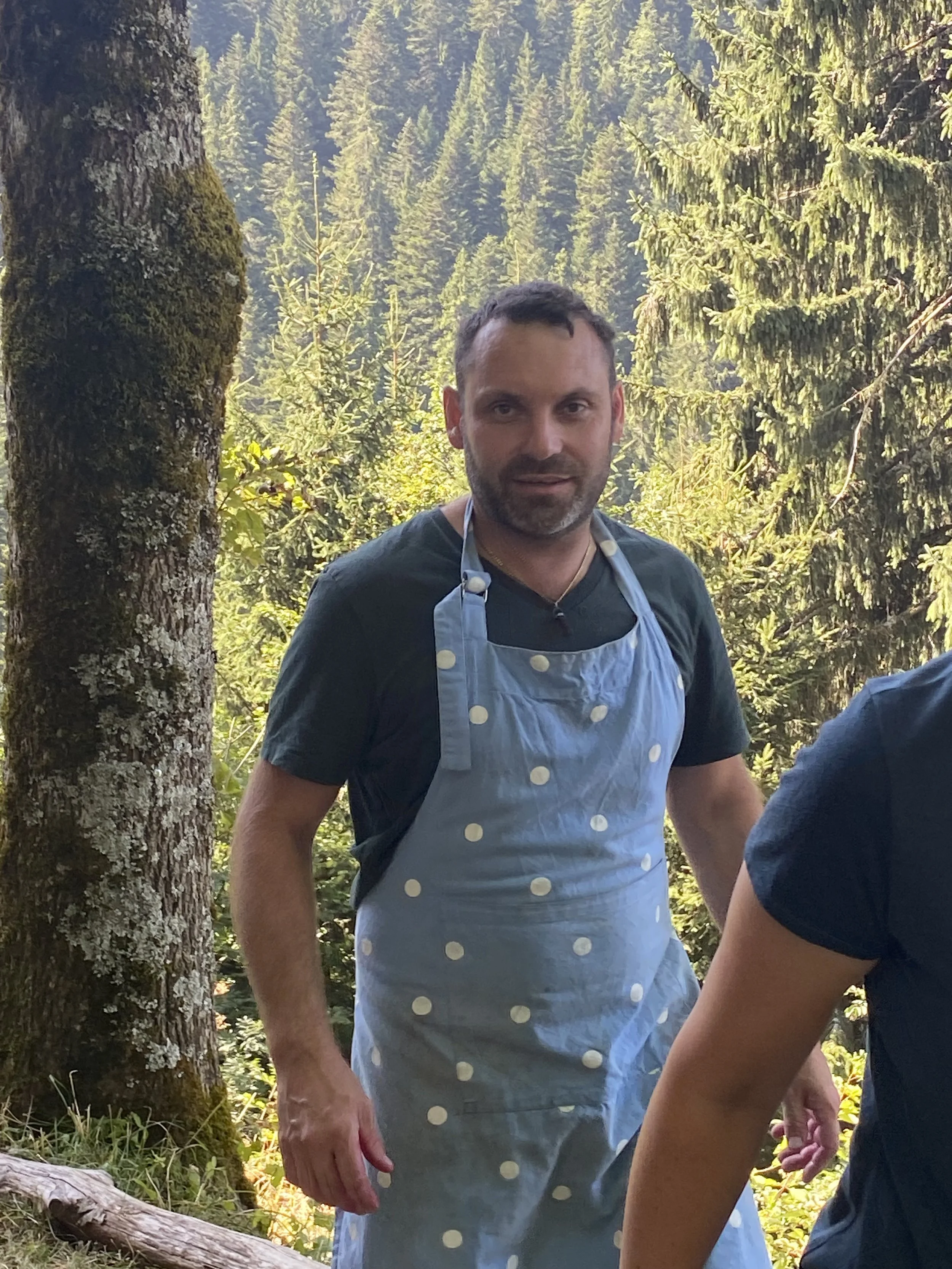 A man wearing a blue polka dot apron stands outdoors in a forested area with tall trees and dense greenery in the background.